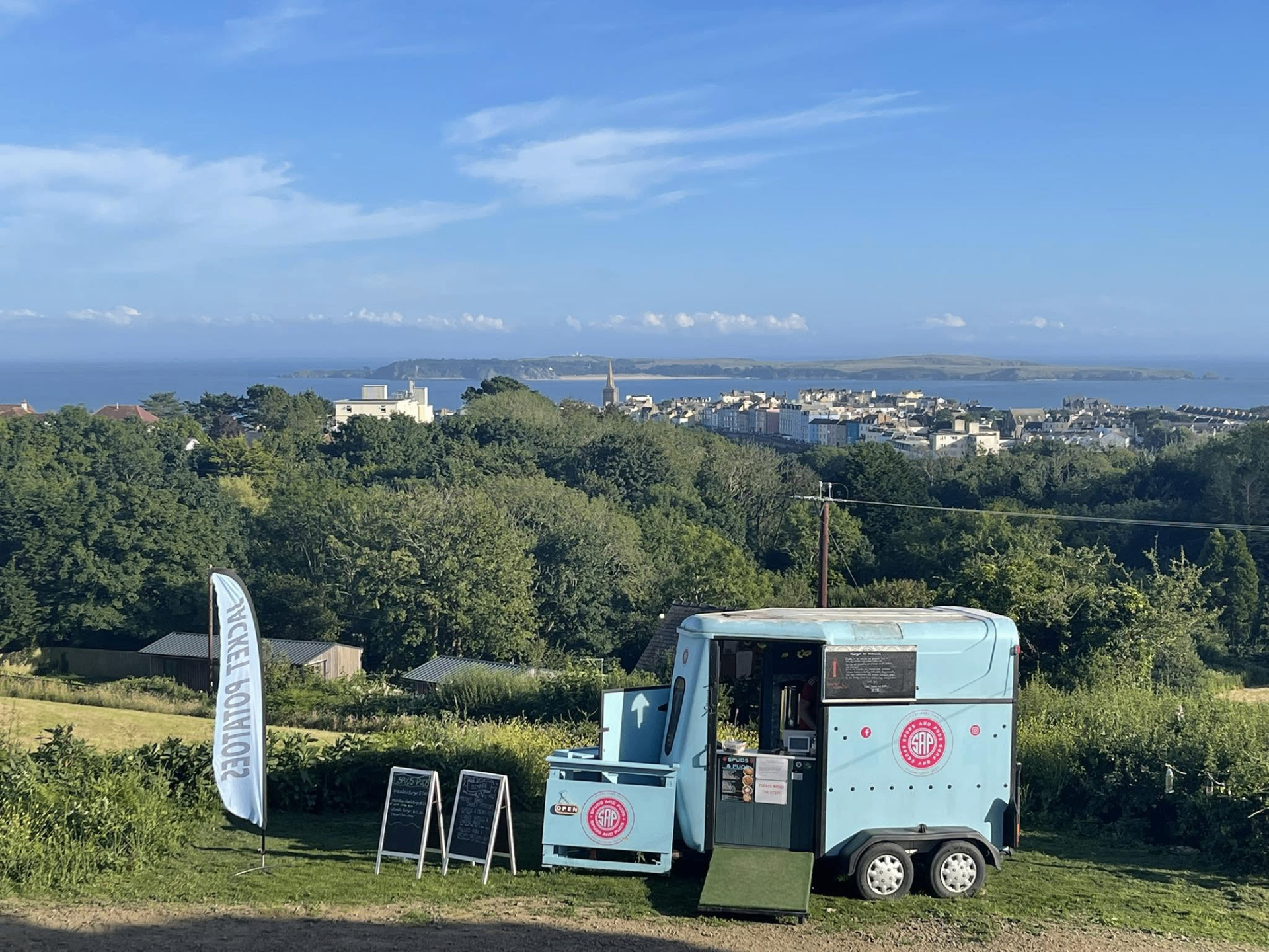 The Spuds & Puds pale-blue converted horsebox trailer parked above Tenby with the coast in the background